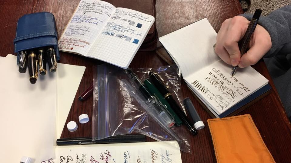 A top-down view of a table where open notebooks displaying cursive writing in ink are visible, and a hand holding a fountain pen is writing in one notebook.  Pen holders with pens are also visible.