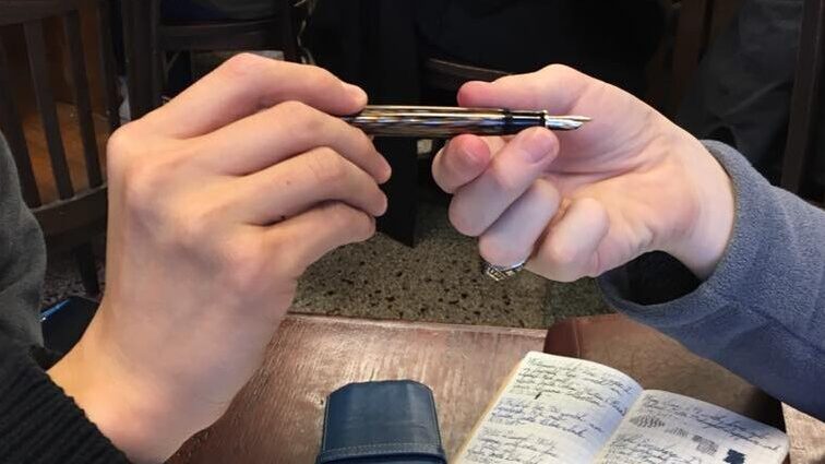 An image of one hand passing a open fountain pen to another hand.  A table with an open notebook is in the background