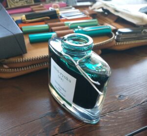 an open glass bottle of teal ink on a wooden table, with fountain pens in a case in the background
