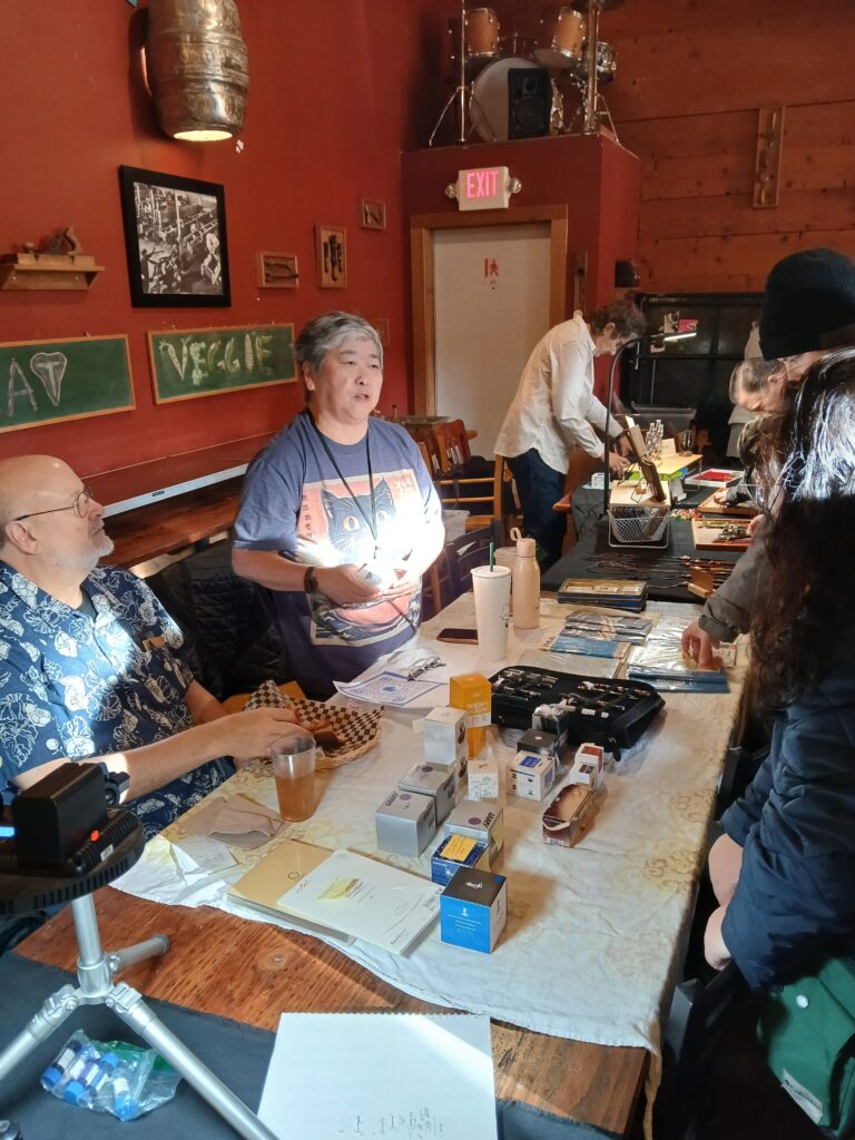 A man and a woman behind a table talking with a customer. Bottles of ink are on the table.